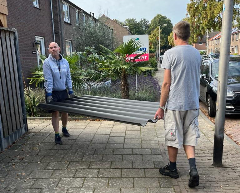 Twee personen hebben een stuk golfplaat vast en tillen deze door de voortuin.