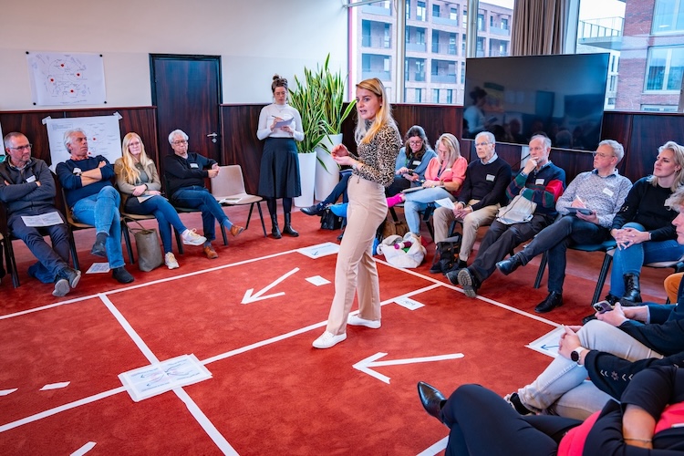 Een vergaderzaal met pijlen op de grond. Iemand staat in het midden en er zit een grote groep mensen in een cirkel eromheen.