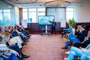 Een vergaderzaal vol met mensen, waar een spreker een presentatie geeft voor een groot scherm.