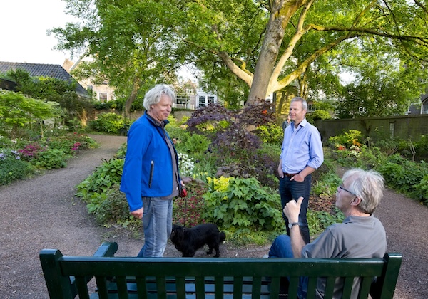 Drie mensen in een hond maken een praatje met elkaar in een parkje. Eén iemand zit op een bankje en de andere twee personen staan voor het bankje.