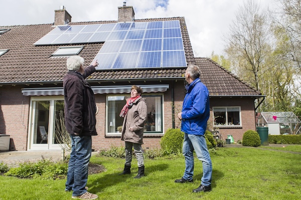Drie mensen staan voor een huis. Eén van de personen wijst naar de zonnepanelen die op het dak van het huis liggen.