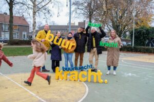 Een groep mensen staat lachend op een basketbalveldje in een woonwijk. Ze hebben Buurkracht letters gemaakt van piepschuim vast en kijken vrolijk naar elkaar en naar de camera. Door het beeld rennen nog twee kinderen.