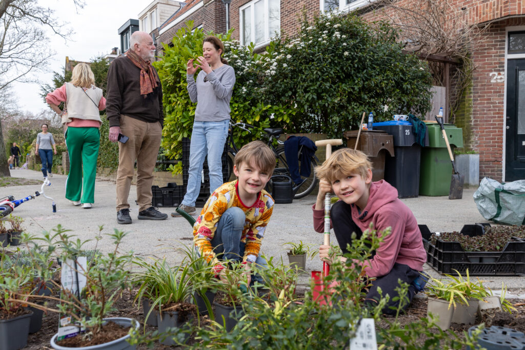 Twee kinderen planten in Leiden samen met hun buren boomspiegels, ze kijken lachend naar de camera.